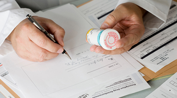 A close-up of a male doctor's hands writing a prescription while inspecting the label of a bottle of medication, illustrating how behavioural science can impact healthcare provider behaviour