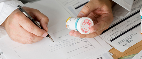 A close-up of a male doctor's hands writing a prescription while inspecting the label of a bottle of medication, illustrating how behavioural science can impact healthcare provider behaviour
